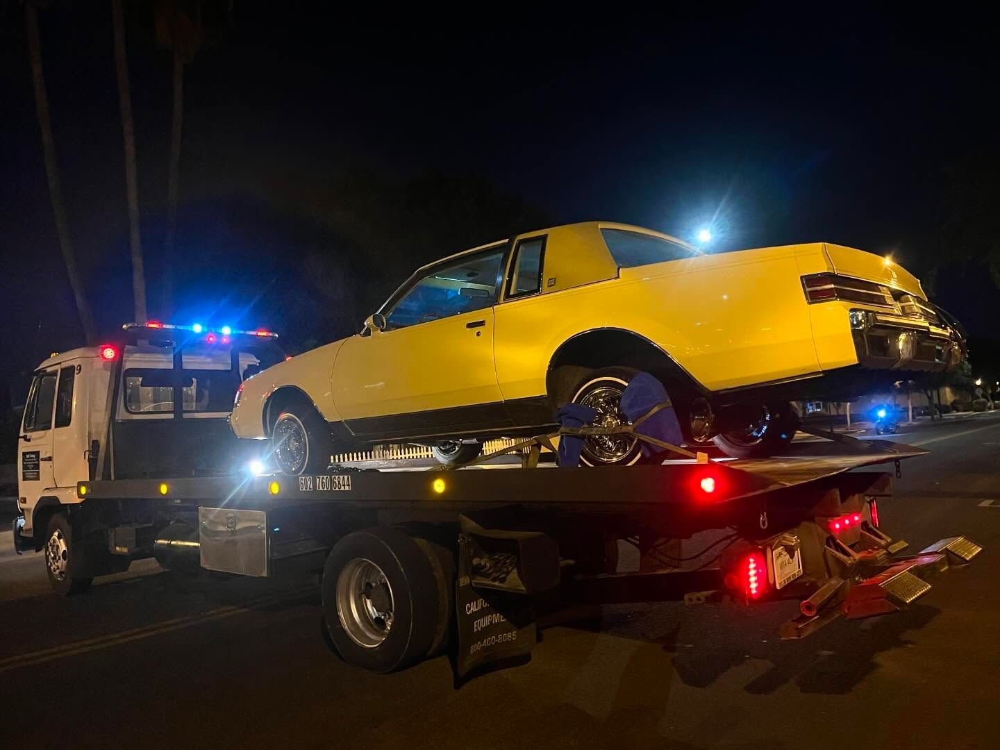 A yellow Buick Regal being towed by a tow truck at night. The truck has flashing lights.