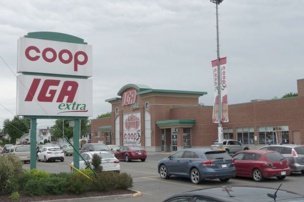 Extérieur de l'épicerie Coop IGA Extra avec des voitures garées devant par une journée nuageuse.
