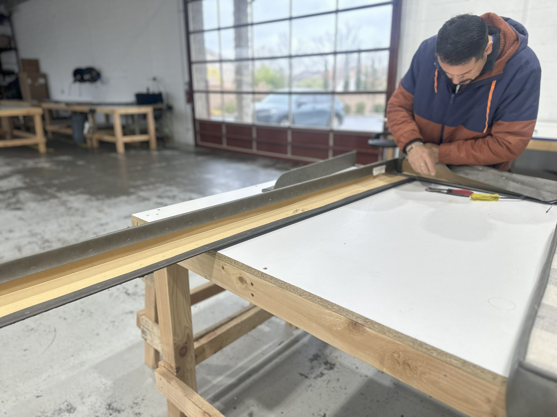 People working in a workshop; cutting, assembling materials. Interior shot, tools, equipment, and materials visible.