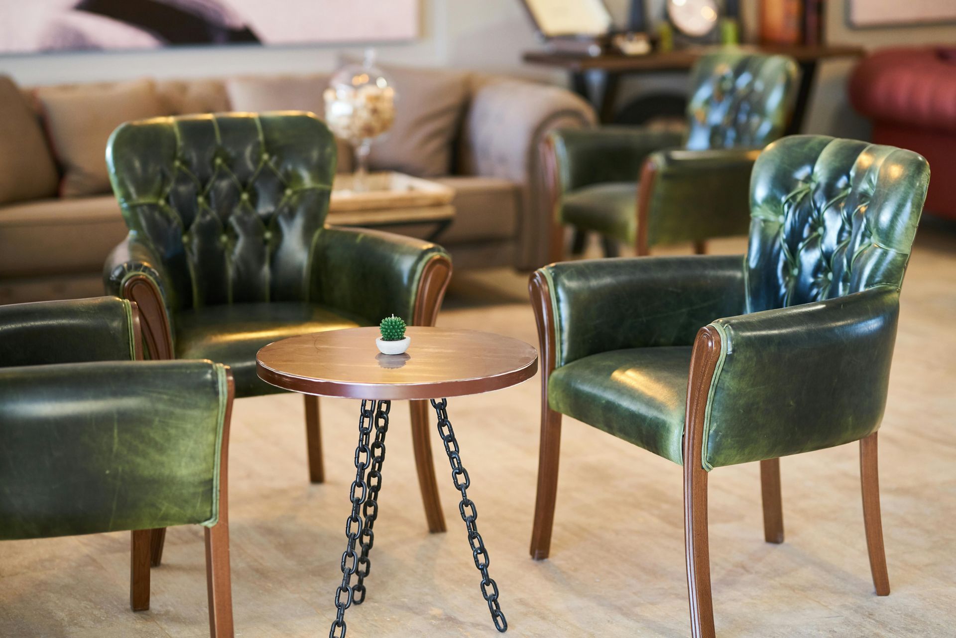 A seating area with four green leather armchairs around a small, round wooden table. A sofa is in the background.