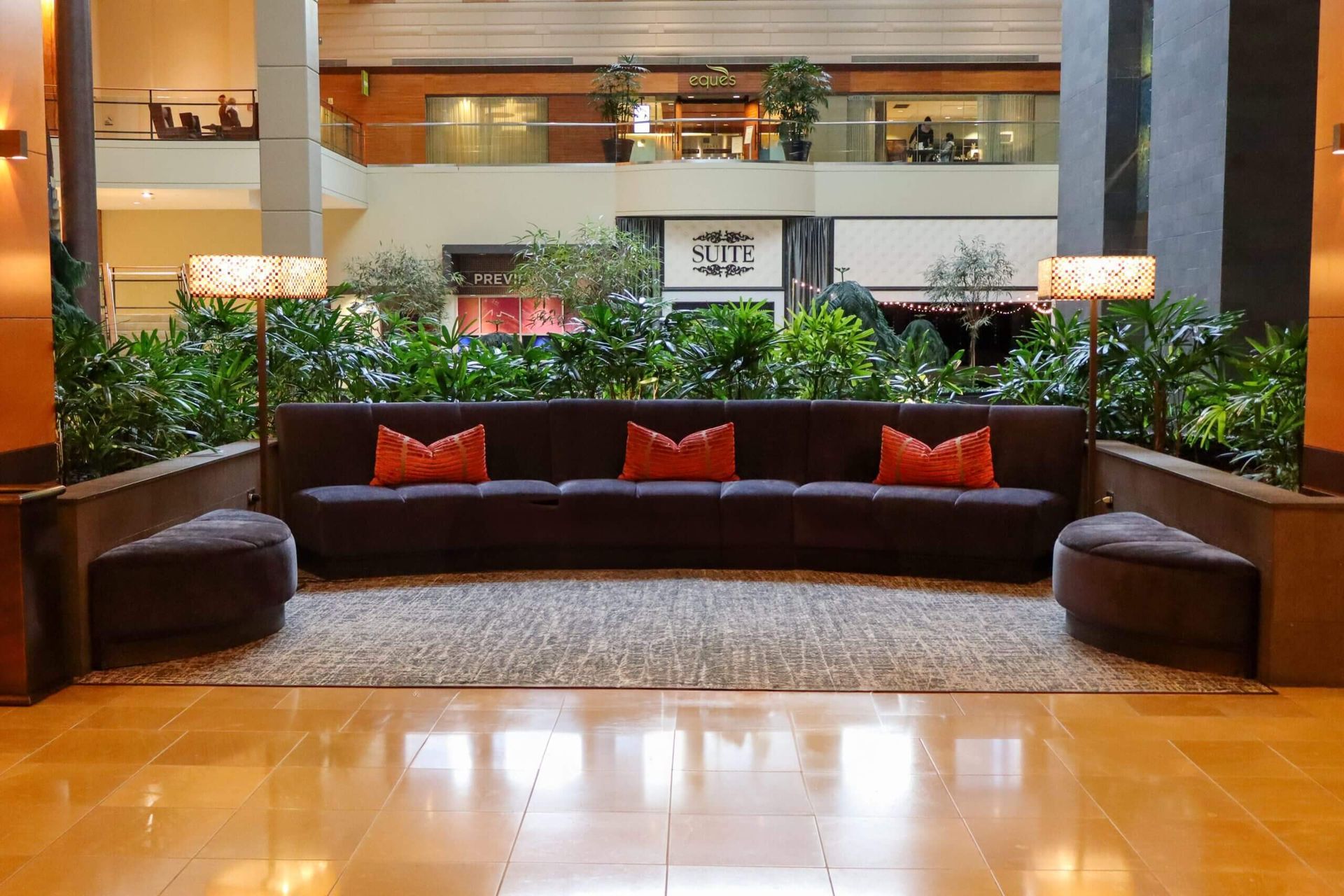 Lobby seating area with a long dark couch, orange pillows, plants, lamps, and a patterned rug on a polished floor.