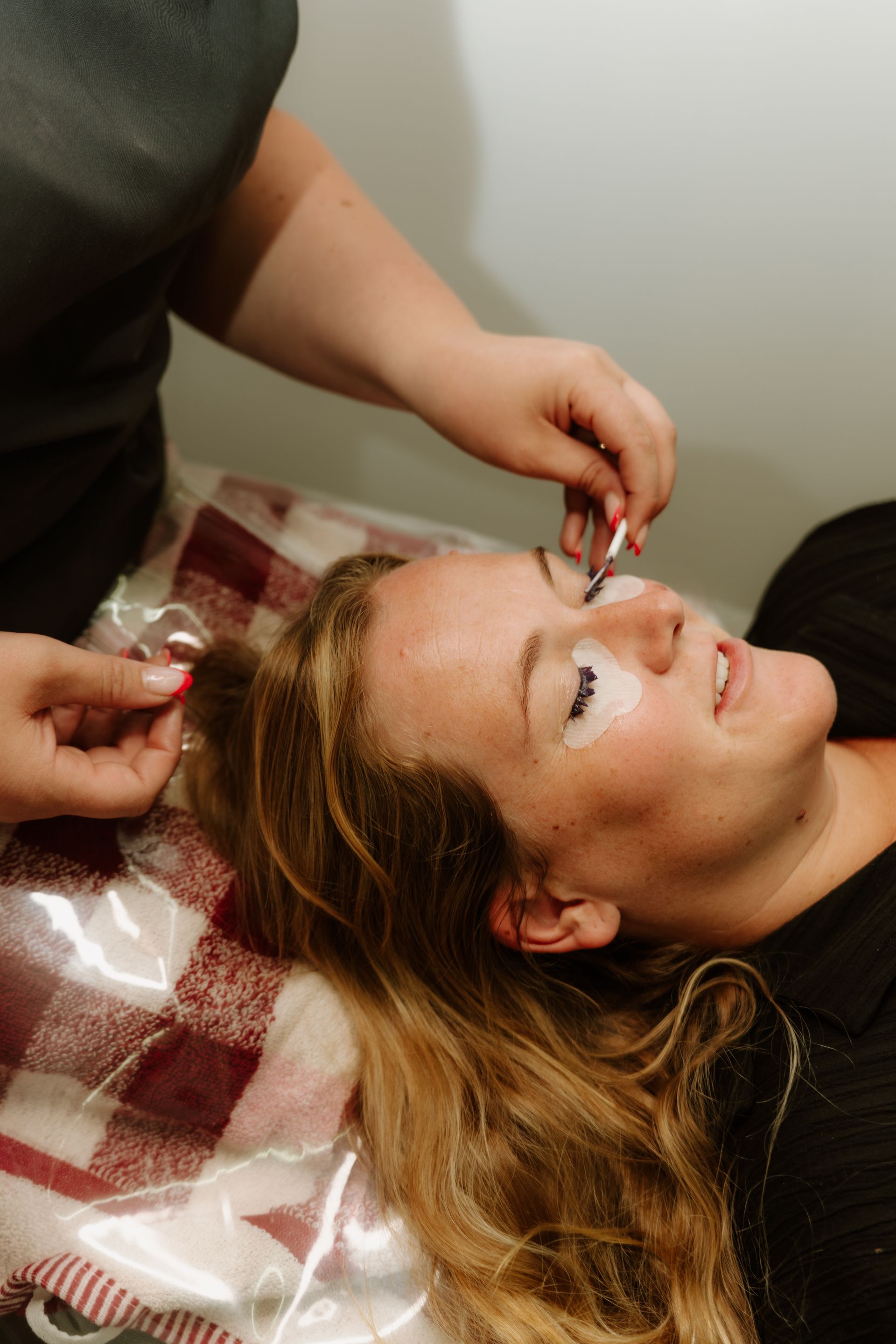 Woman receiving eyelash extensions; technician using tweezers. White eye pads, red/white blanket. — Admire Skincare and Beauty in Taree, NSW