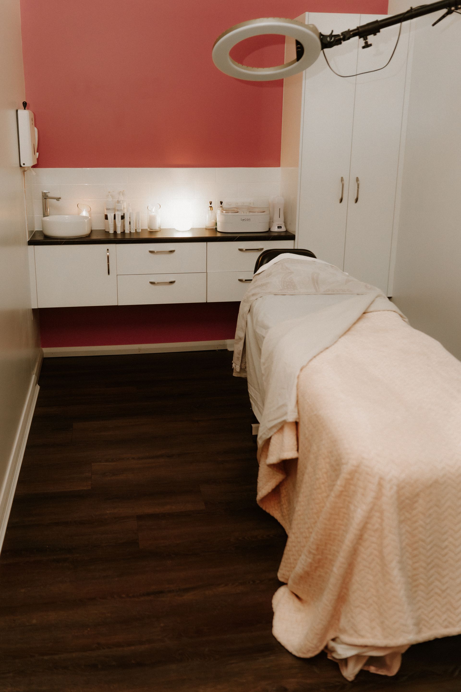 Salon treatment room with a bed covered in light pink linens and cabinets. Dark wood floors.-  — Admire Skincare and Beauty in Taree, NSW
