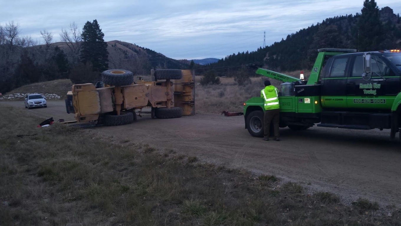 A roadside assistance worker kneels to secure a chain to a car wheel, a tow truck behind.