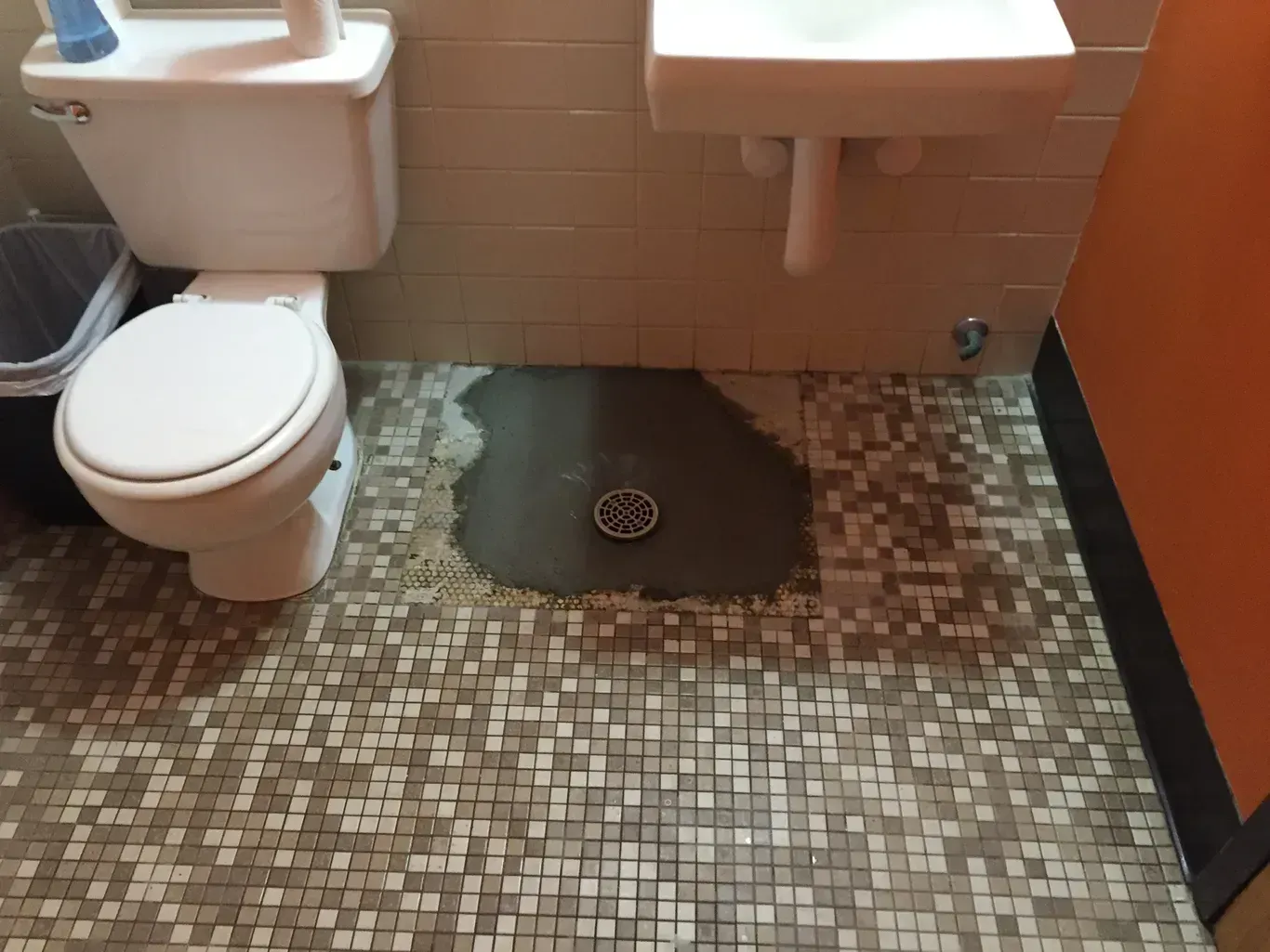 Public restroom with a toilet, sink, and water pooling around a floor drain.