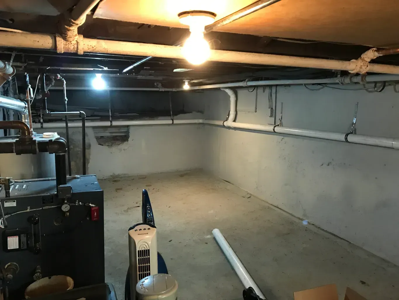 Basement interior with exposed pipes, concrete floor, furnace, and three ceiling lights. White and gray walls.