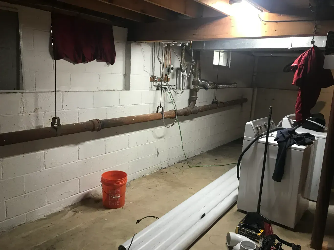 Basement with painted white block walls, pipes, a washing machine, and a red bucket.