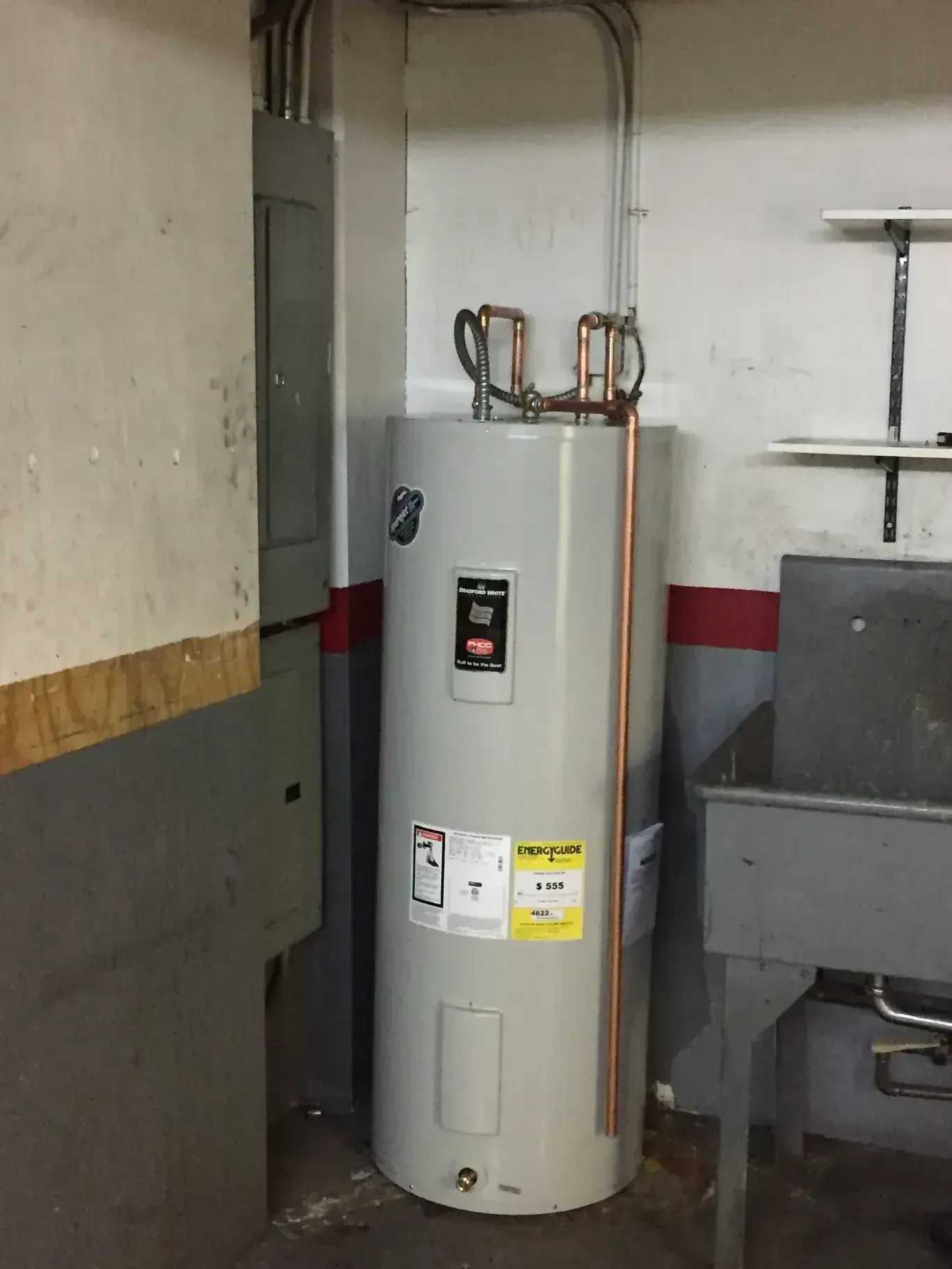 Tall, gray water heater in a basement corner with copper pipes and electrical wiring.
