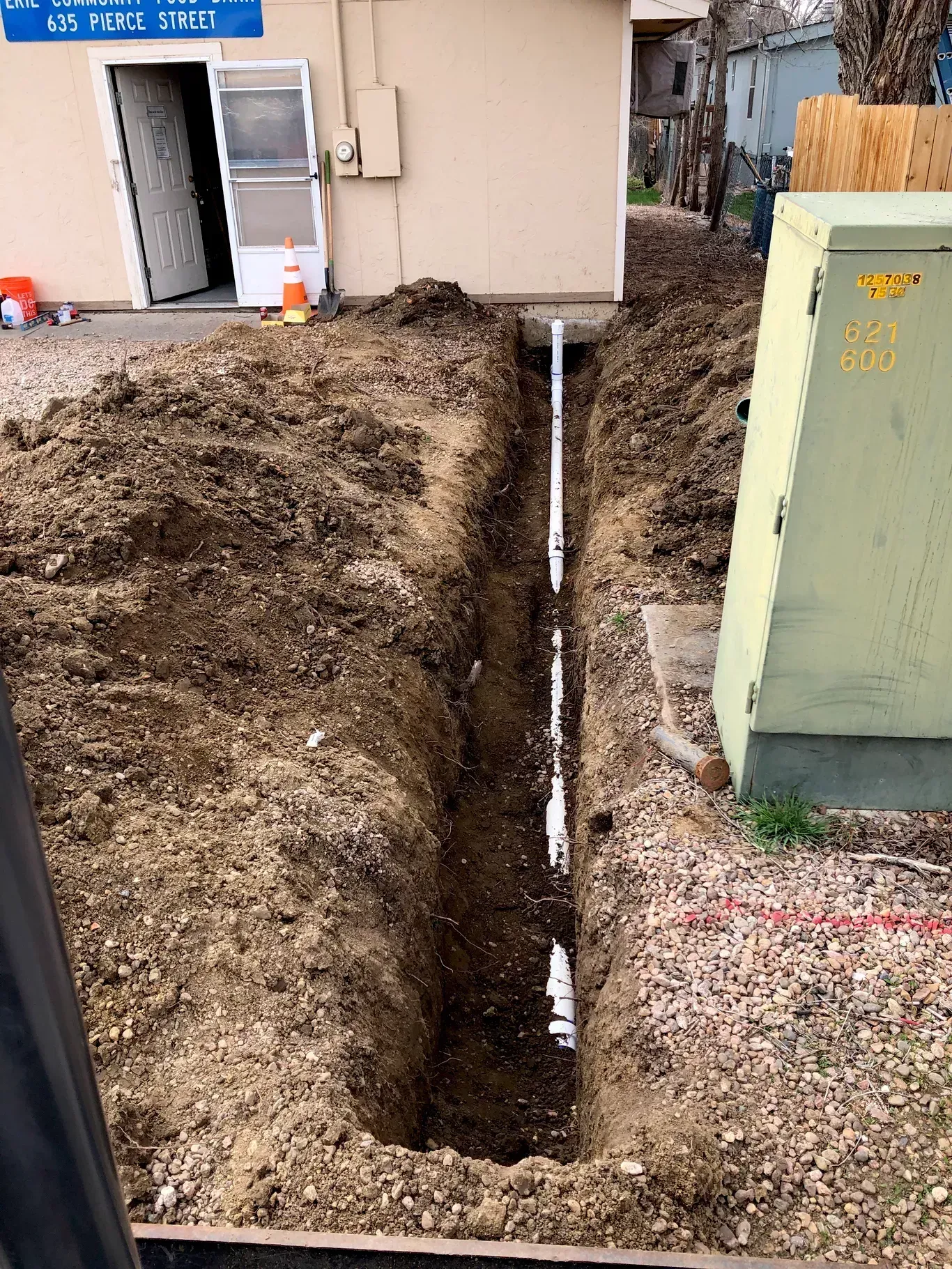 A trench dug next to a building, containing a white pipe. A utility box is on the right.