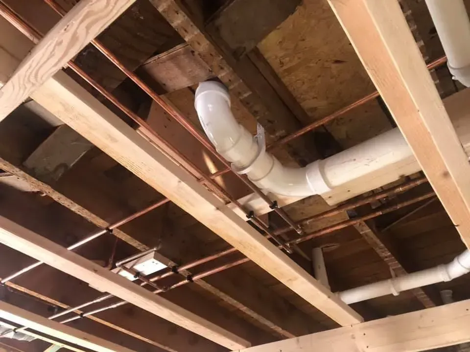 Plumbing pipes (copper and PVC) and wooden support beams visible beneath a floor.