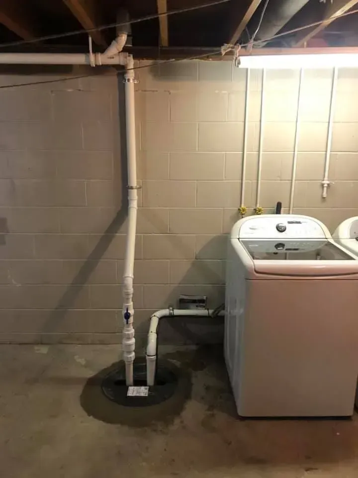 A white washing machine next to a sump pump system in a basement with gray cinder block walls.