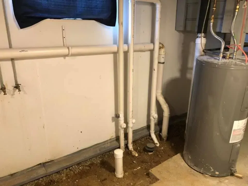 Basement with pipes, water heater, and drain. White walls and pipes, gray water heater, brown floor.