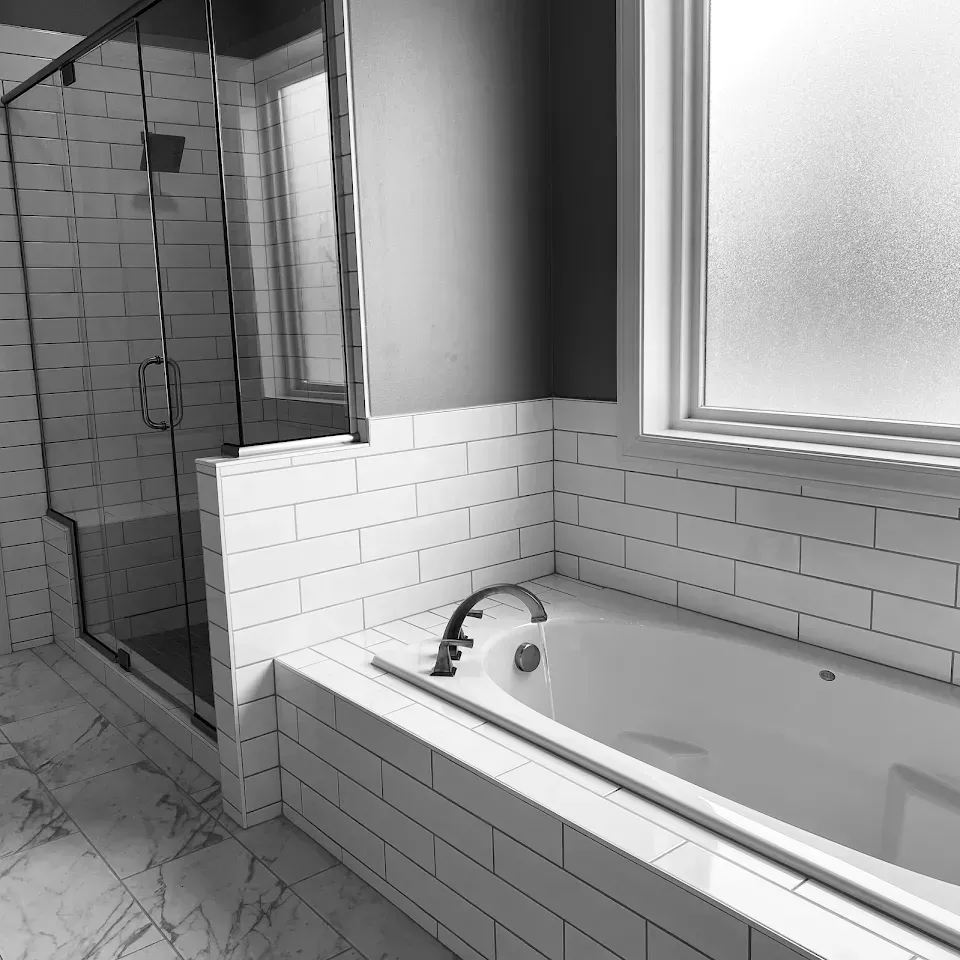 Black and white photo of a modern bathroom with a tub, shower, and window. White subway tile lines the walls.