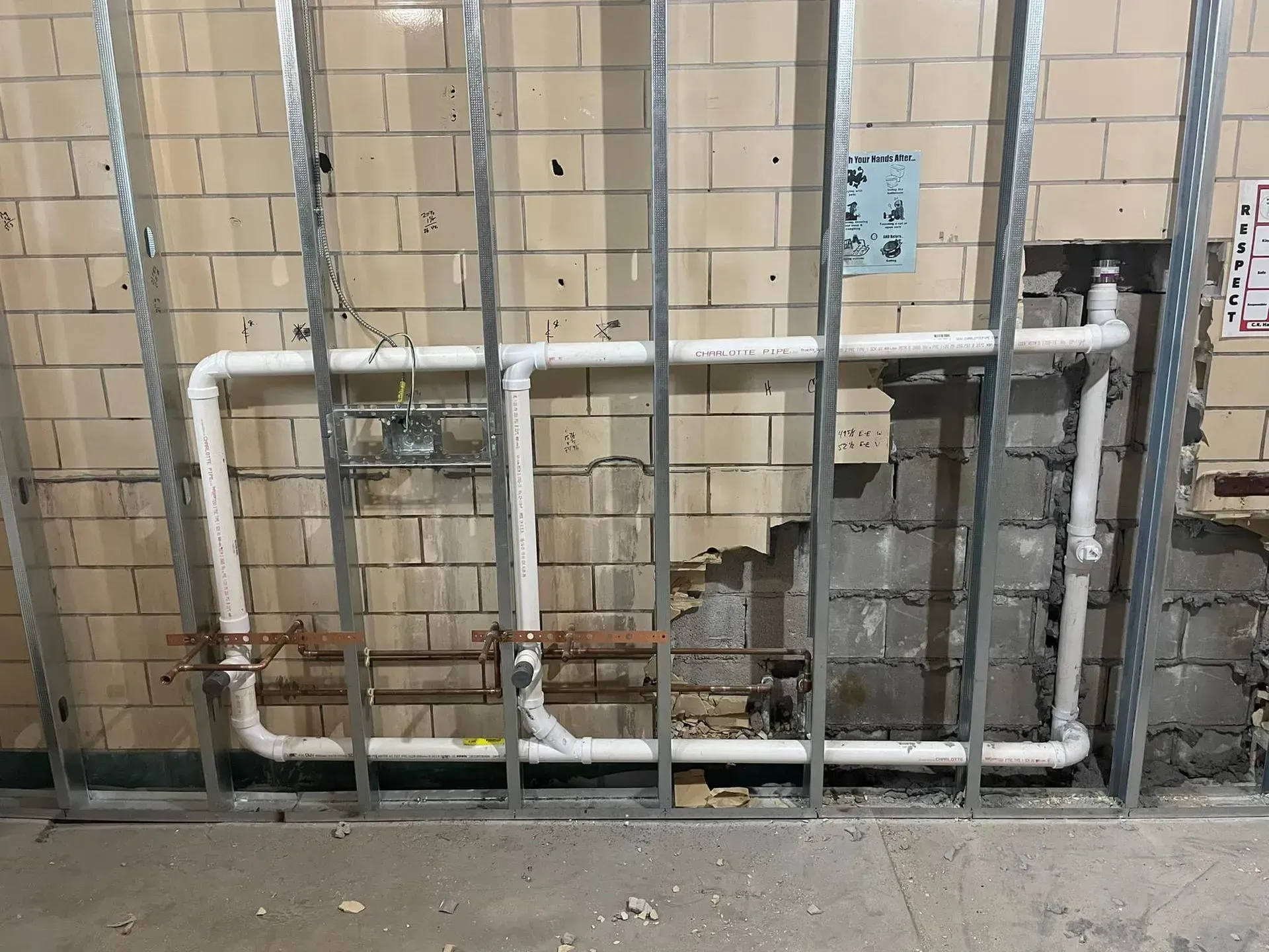 Metal studs frame a wall with plumbing pipes. Damaged brick is visible behind the pipes.