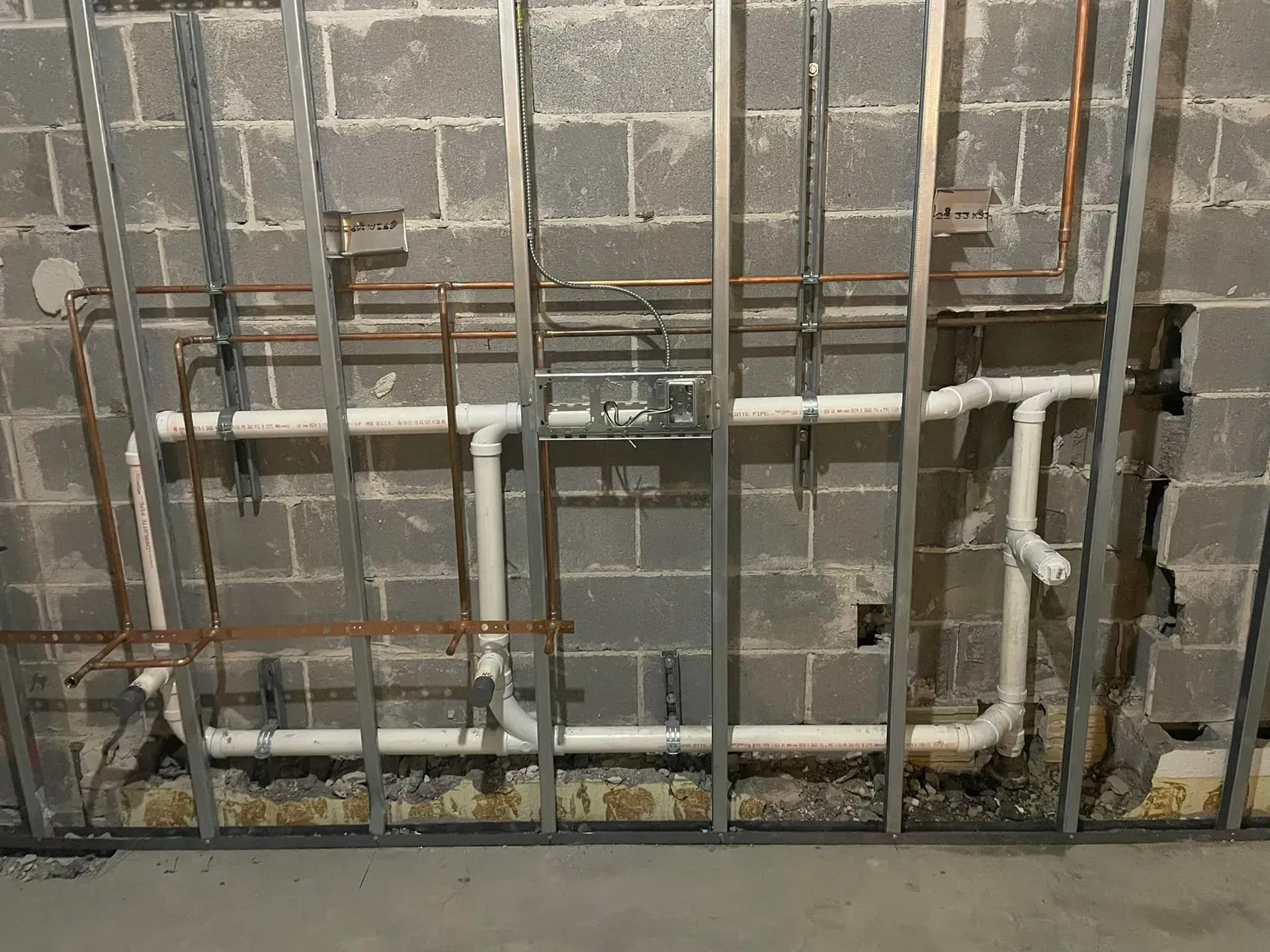 Metal framing with exposed pipes and electrical box against a cinder block wall.