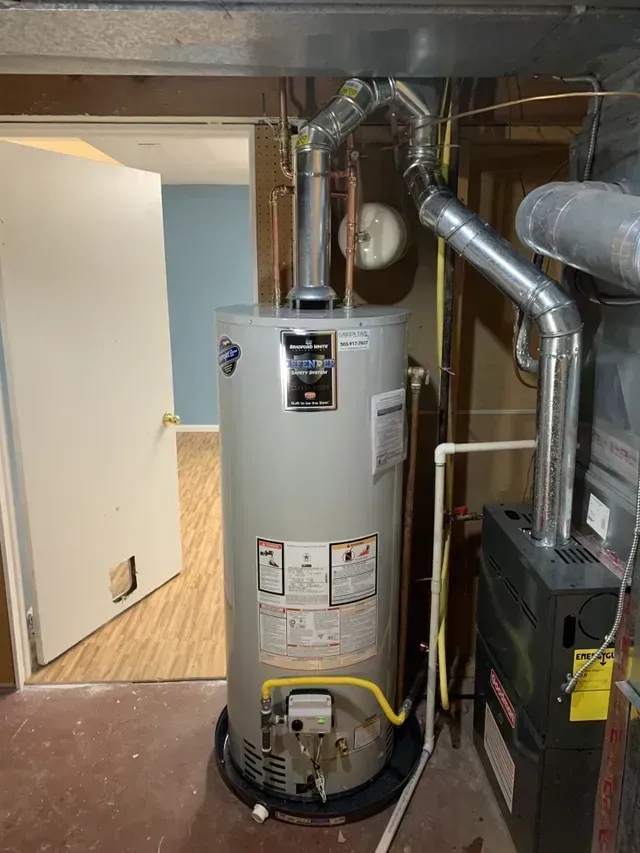 A water heater in a basement with silver venting, and a furnace to its right.