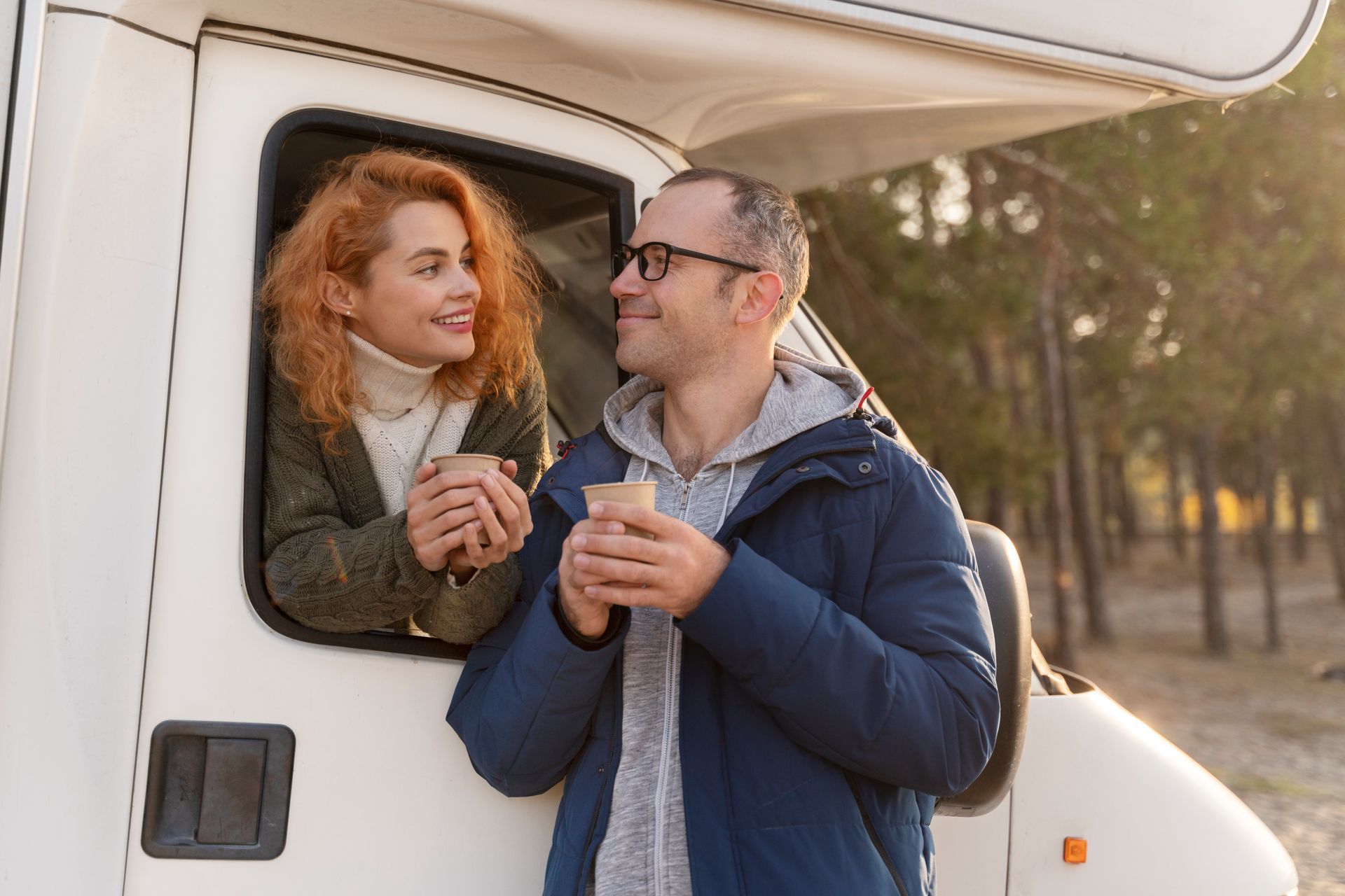 A man and a woman are standing next to a camper van holding cups of coffee.