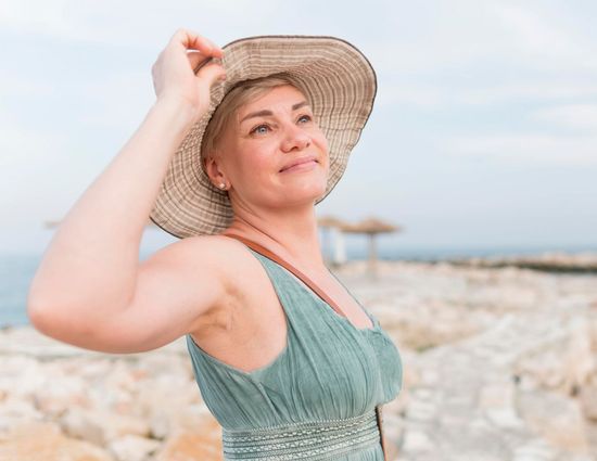 A woman wearing a straw hat and a blue dress is standing on a beach.