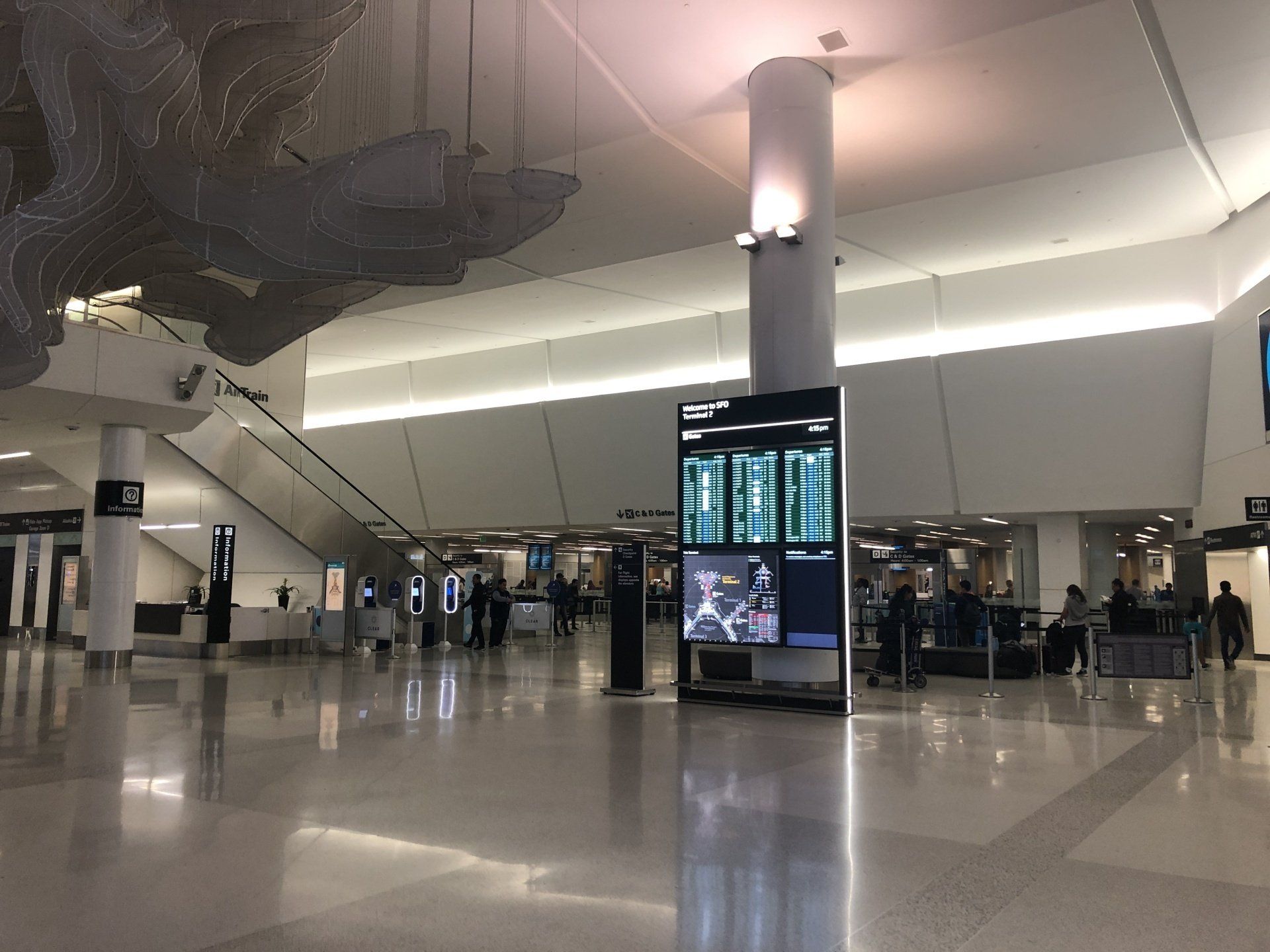 In the center of the photo, a lit wayfinding sign at the terminal in the SFO airport, stretching up to the high ceiling. On the left, an intricate scuplture hangs from the ceiling over the open and empty walkway.