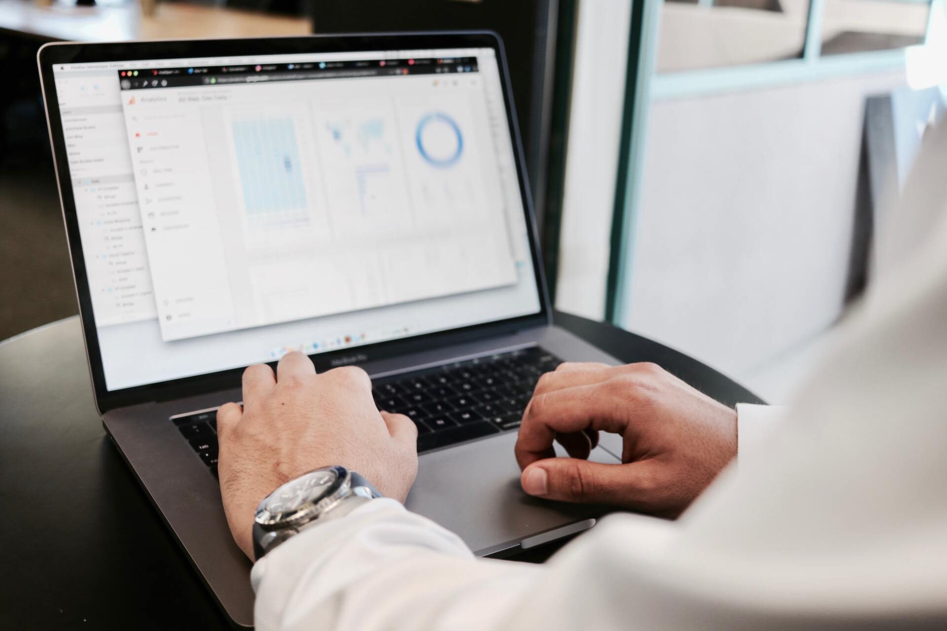 A man's hands and wrists in a dress shirt and wearing a wristwatch rest on an open laptop displaying blurred data and spreadsheets. Pic Campaign Creators via Unsplash.