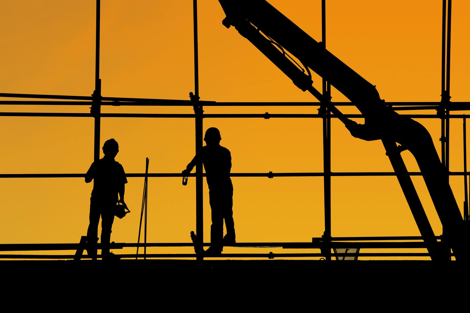 The silhouette of two construction workers wearing hard hats is shown against a orange background in a windowed construction scene. Pic Yancy Min via Unsplash.