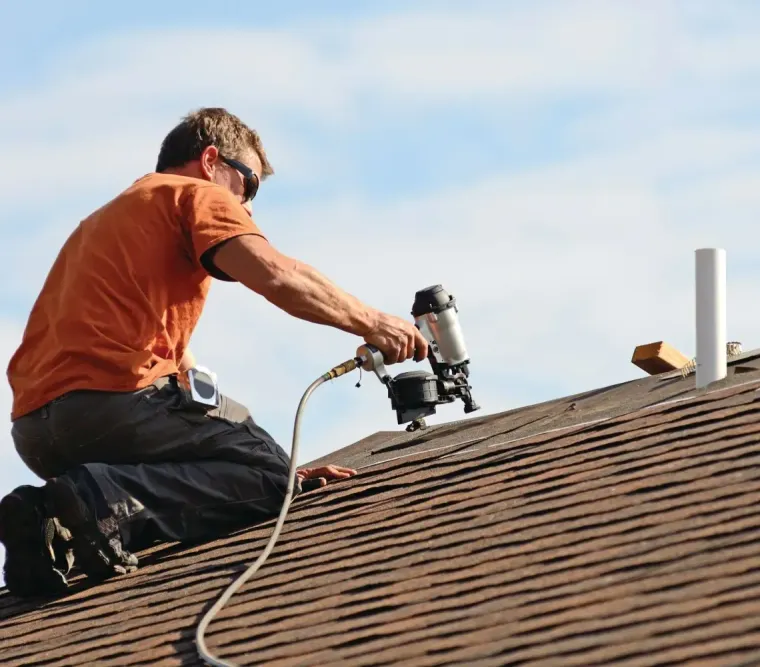 Worker using a power tool to repair a shingled roof under a blue sky