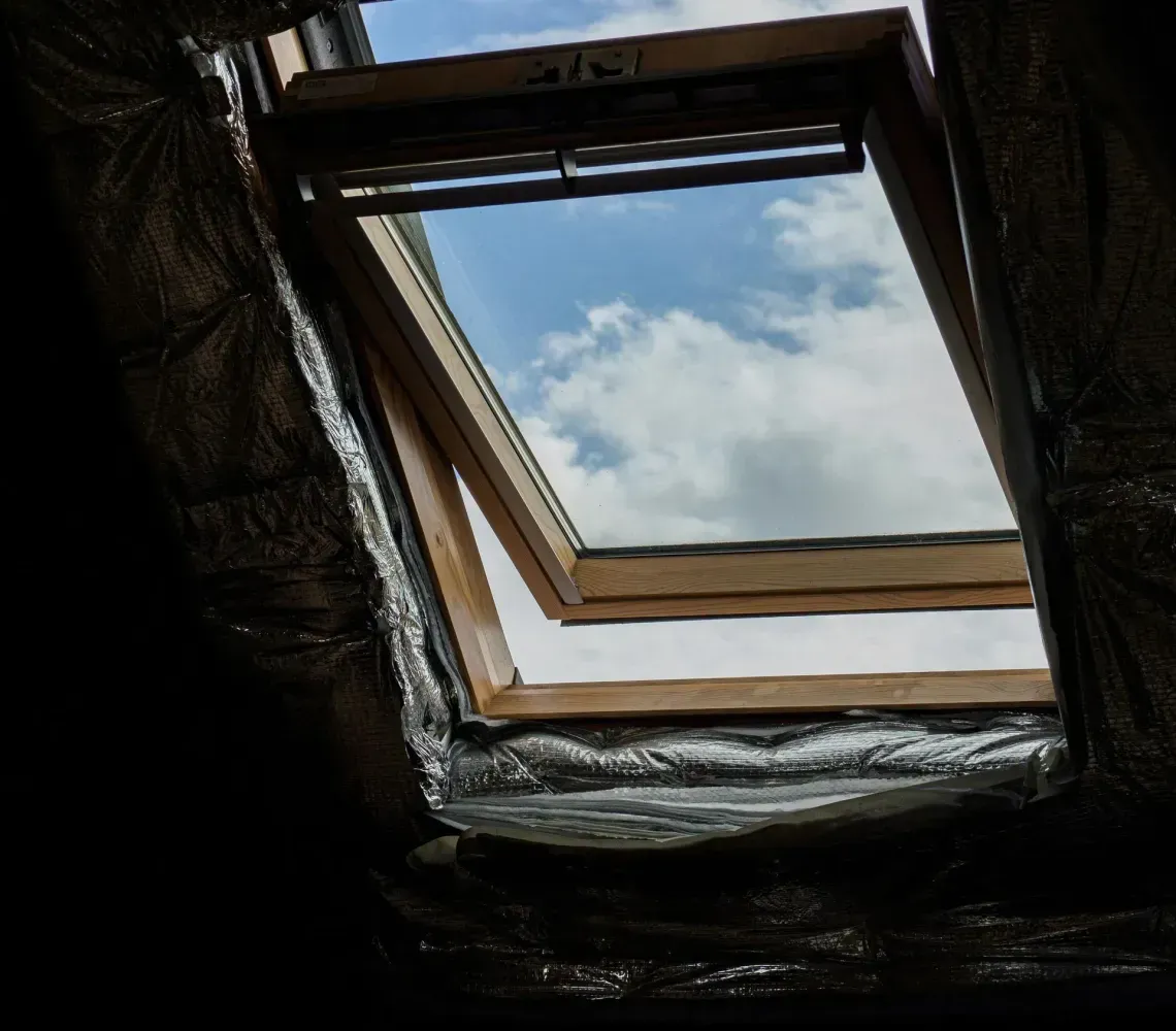 Skylight opening in a dark attic, showing blue sky and clouds above.