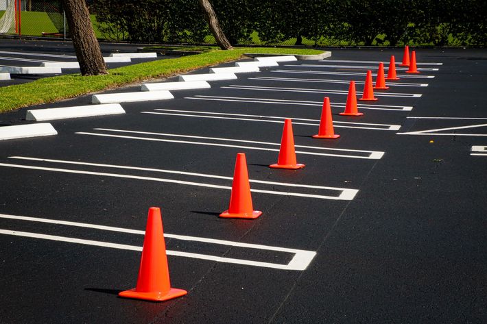 A row of bright orange traffic cones placed along painted parking stall lines in an empty asphalt parking lot.
