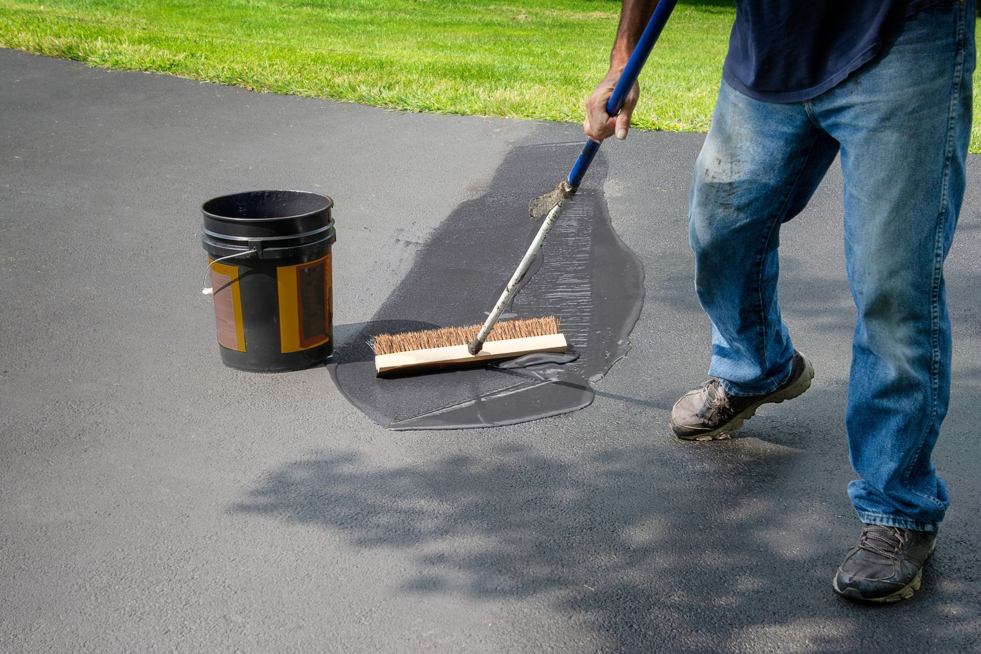 A person uses a brush on a long handle to spread black sealant over cracks on an asphalt driveway near a plastic bucket.