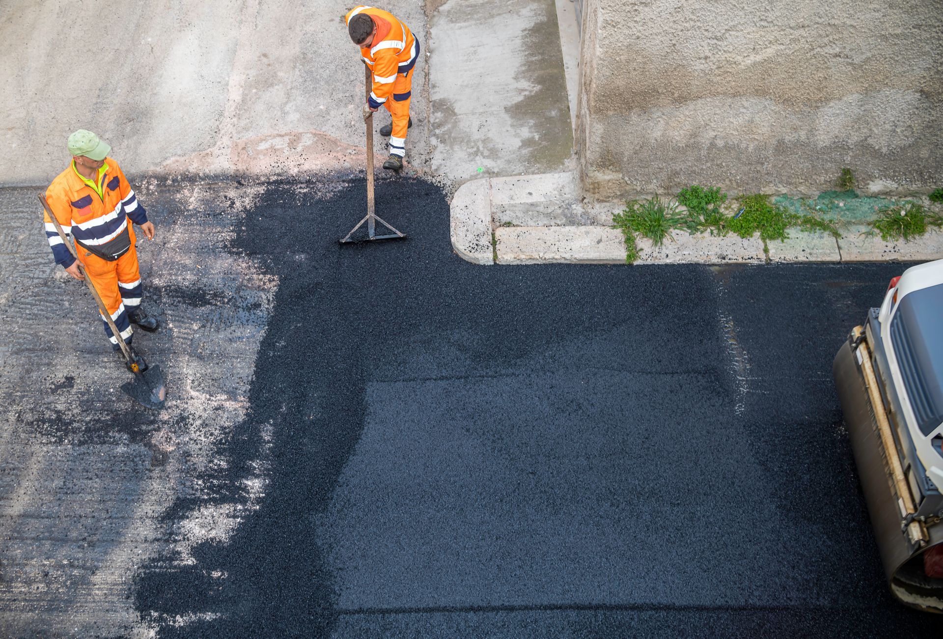 Two road workers in high-visibility orange uniforms smooth freshly laid black asphalt on a street.