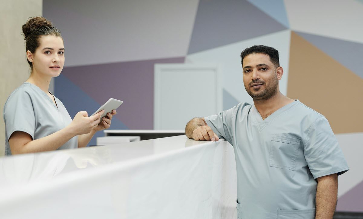 Two medical staff members in light blue scrubs stand behind a reception desk in a modern, geometrically patterned office.