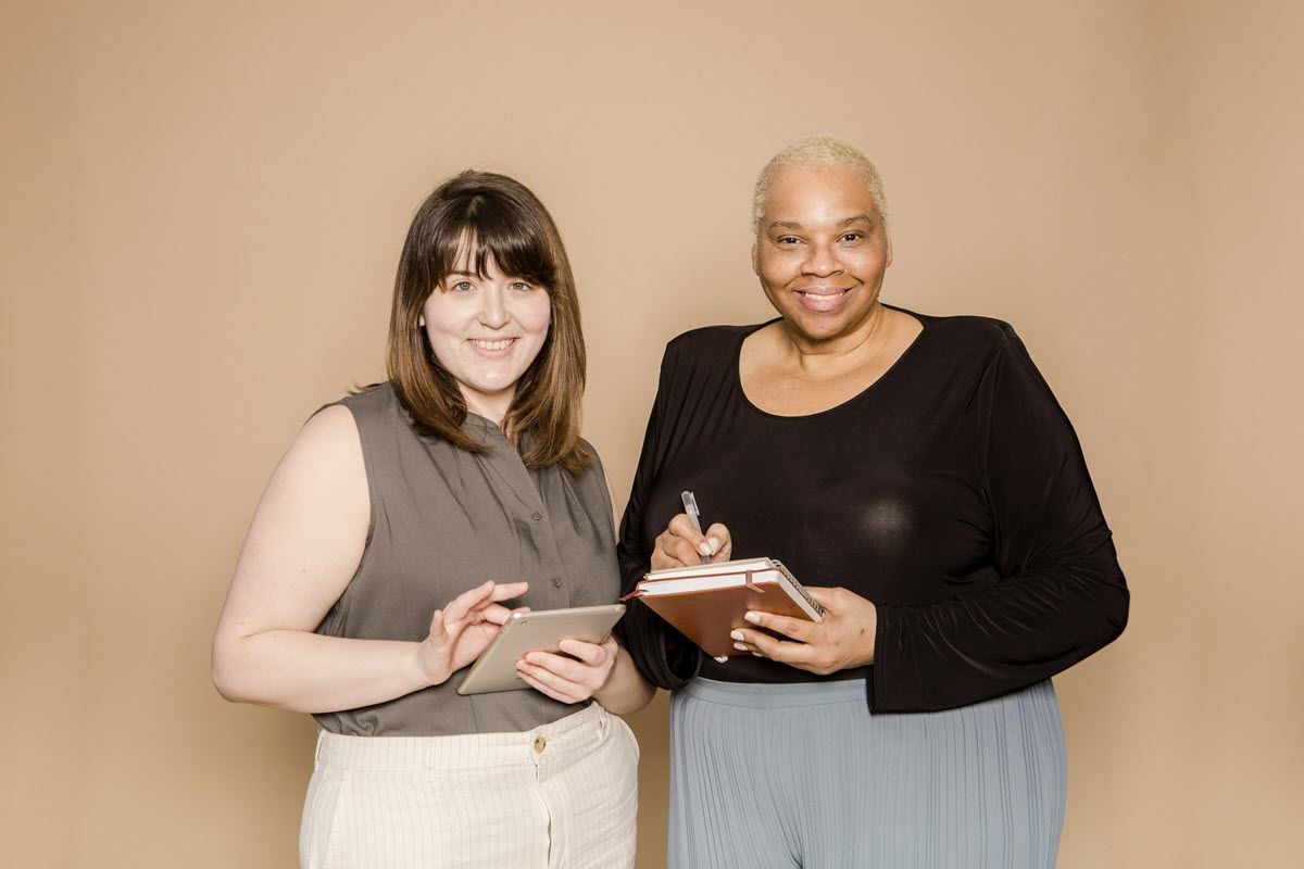 Two women, one with a tablet and one with a notebook, smiling. Plain beige backdrop.