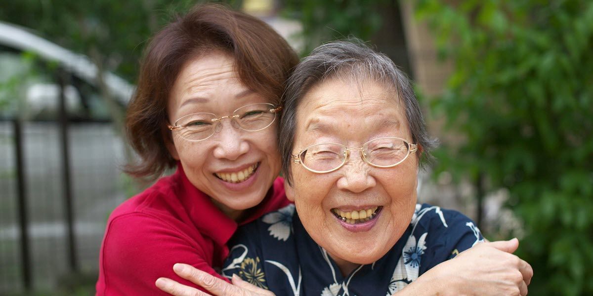 Two smiling women hugging outdoors, one in a red sweater and one in a dark floral shirt.