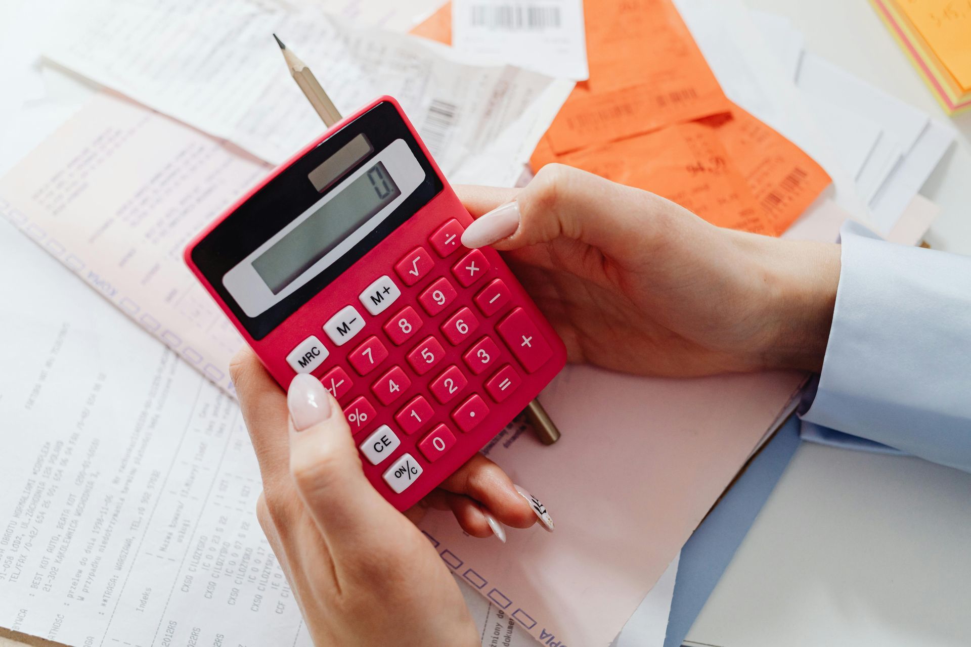 Hands using a red calculator on a desk with paperwork and a pencil.