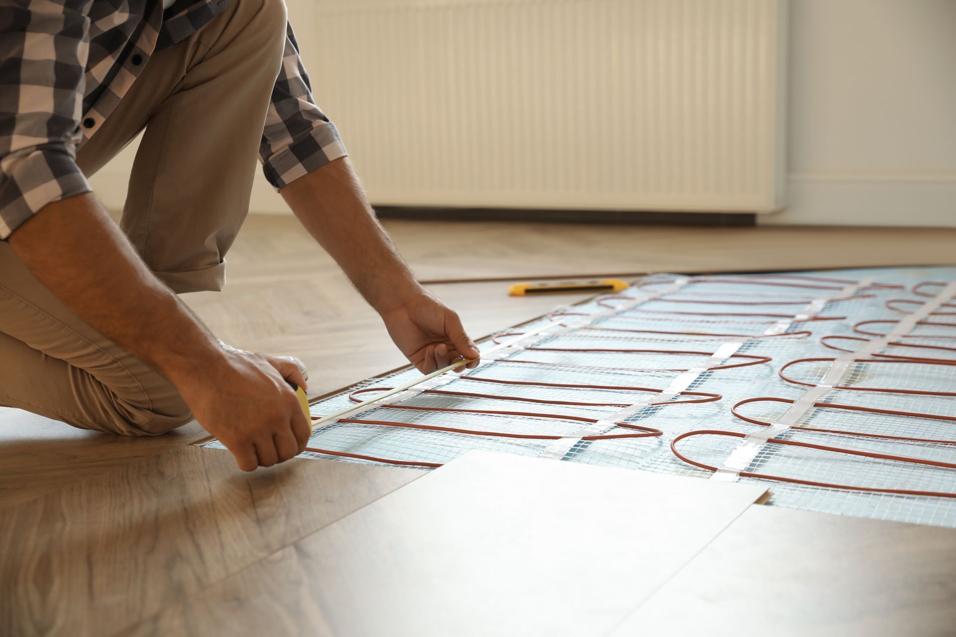 Person installing underfloor heating, measuring against wooden flooring with measuring tape.