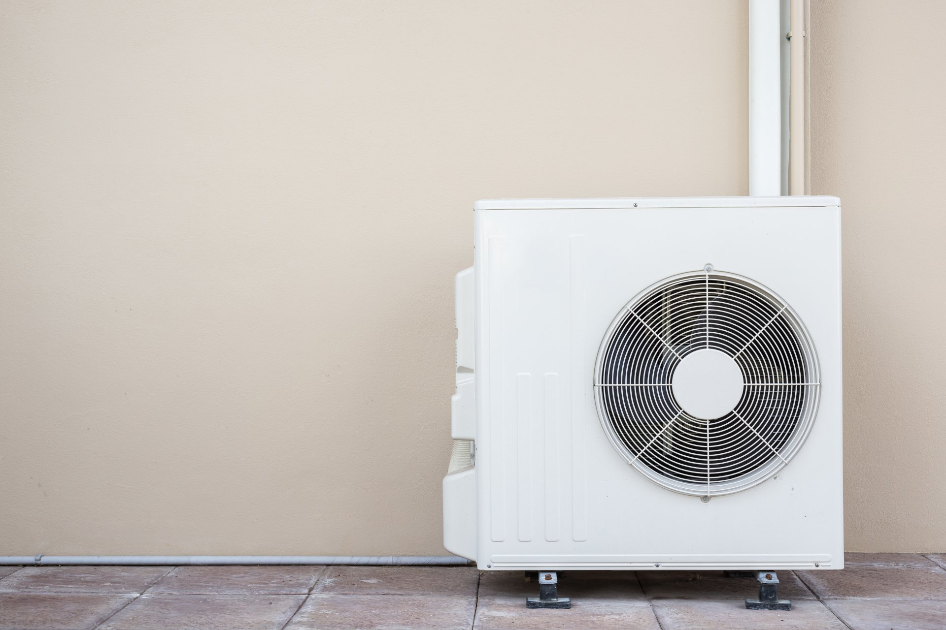 White air conditioning unit outside against a beige wall, pipe visible above.