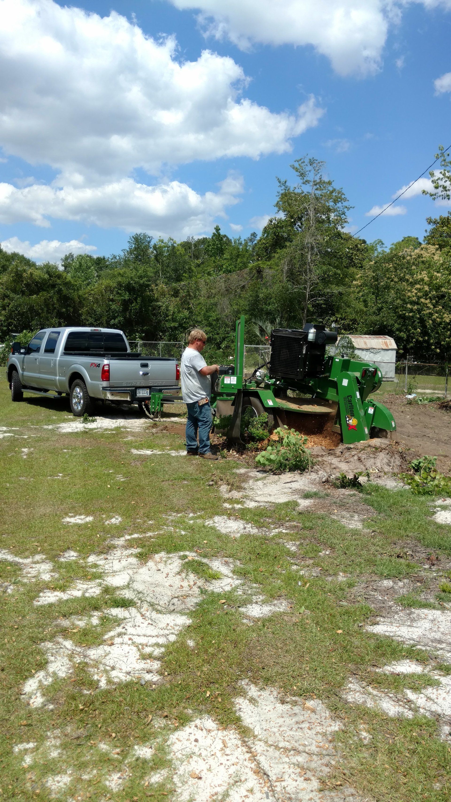 A person operating a stump grinder next to a truck. Blue sky, grassy ground, trees.