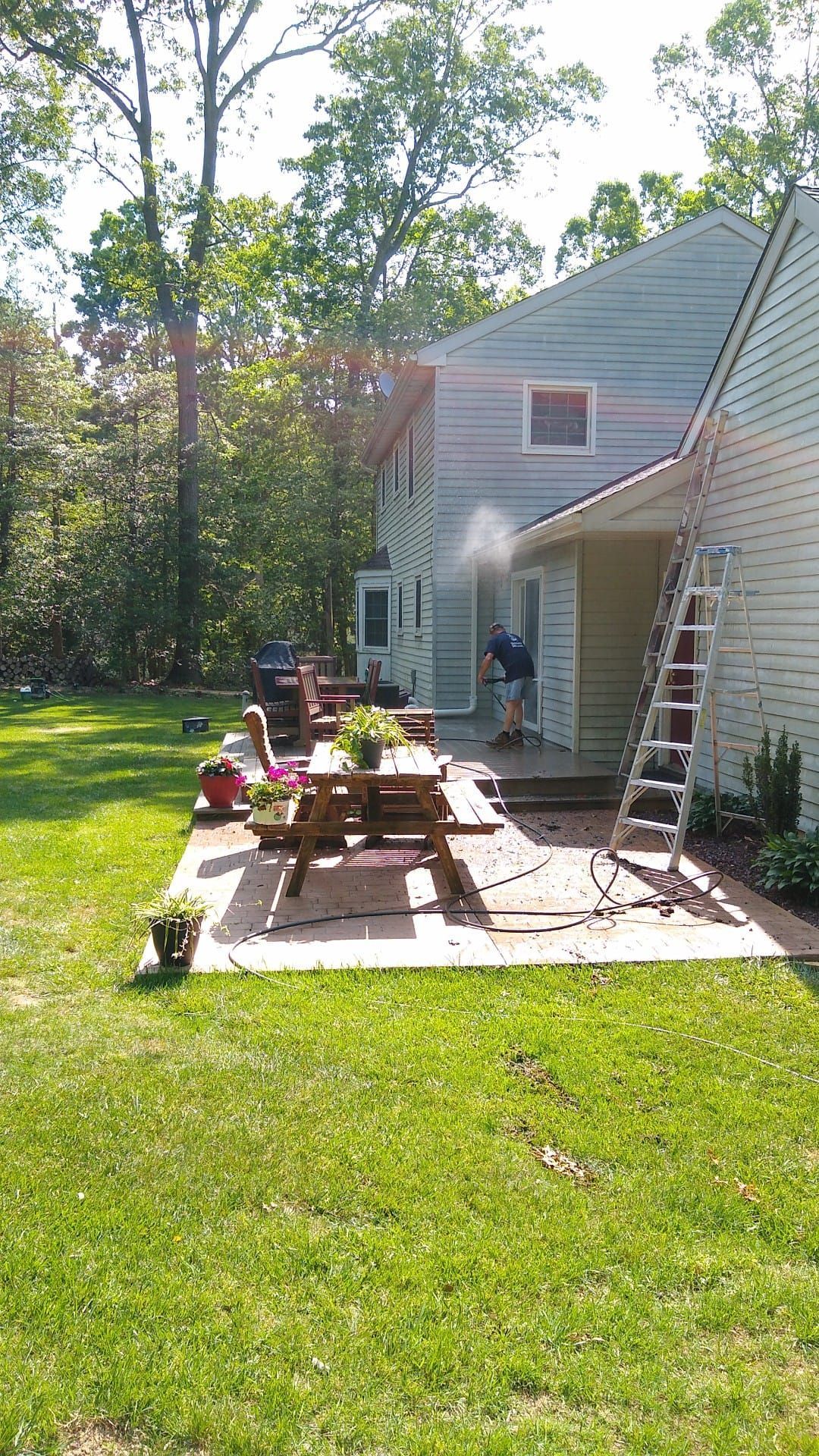 Person pressure washing the side of a house on a sunny day. A picnic table and grill are in the backyard.