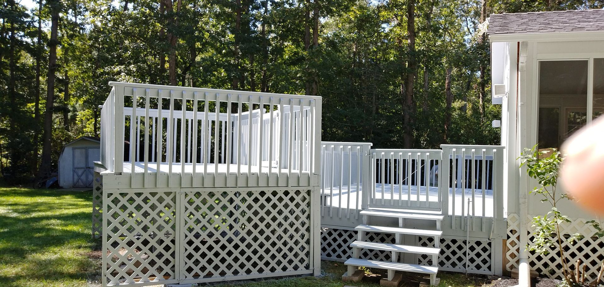 White painted wooden deck with latticework base, steps, and railing, set in a yard with trees.