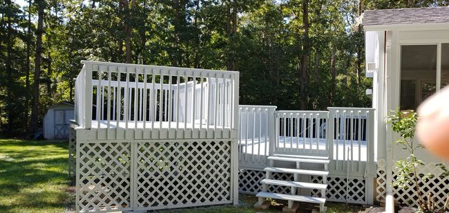 White painted wooden deck with latticework base, steps, and railing, set in a yard with trees.