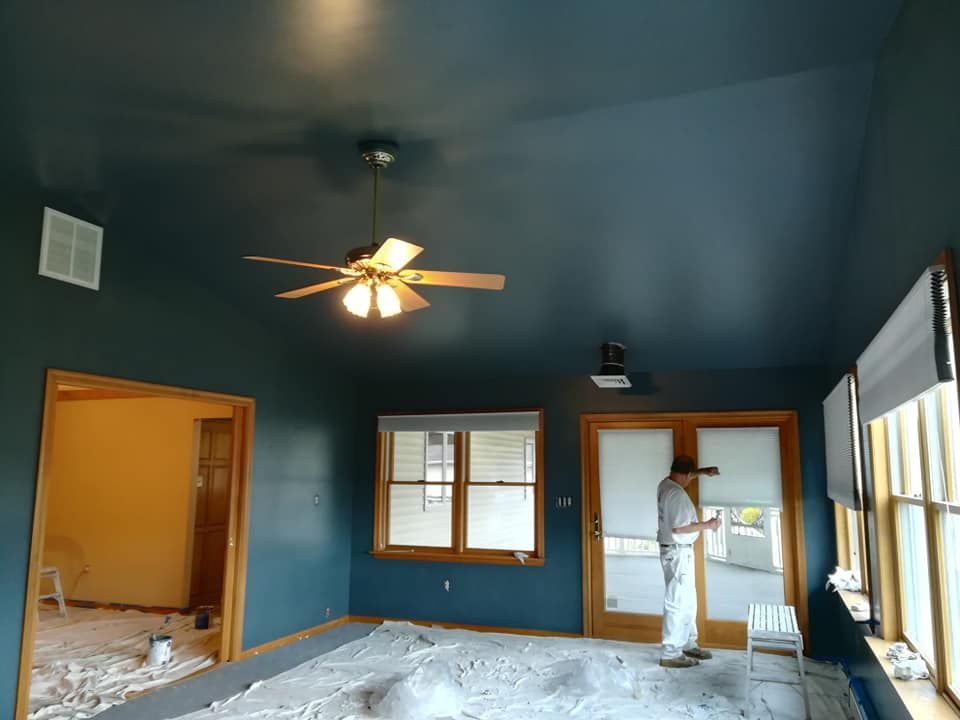 Room with blue walls, ceiling, trim; a person painting. Windows, door, and a ceiling fan are visible.