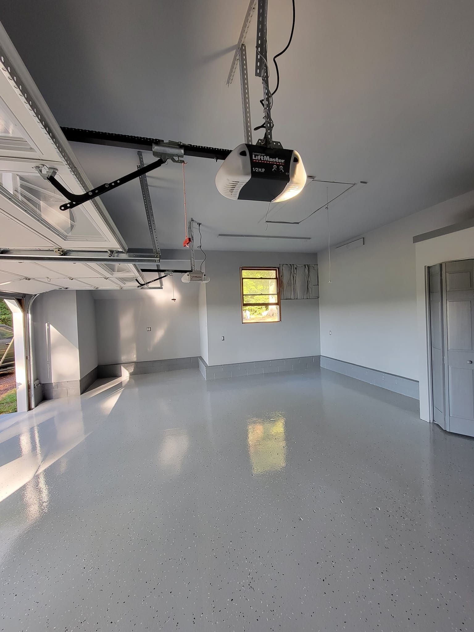 A spacious, newly painted garage with a shiny epoxy floor. A window is in the back wall. The garage door is open.