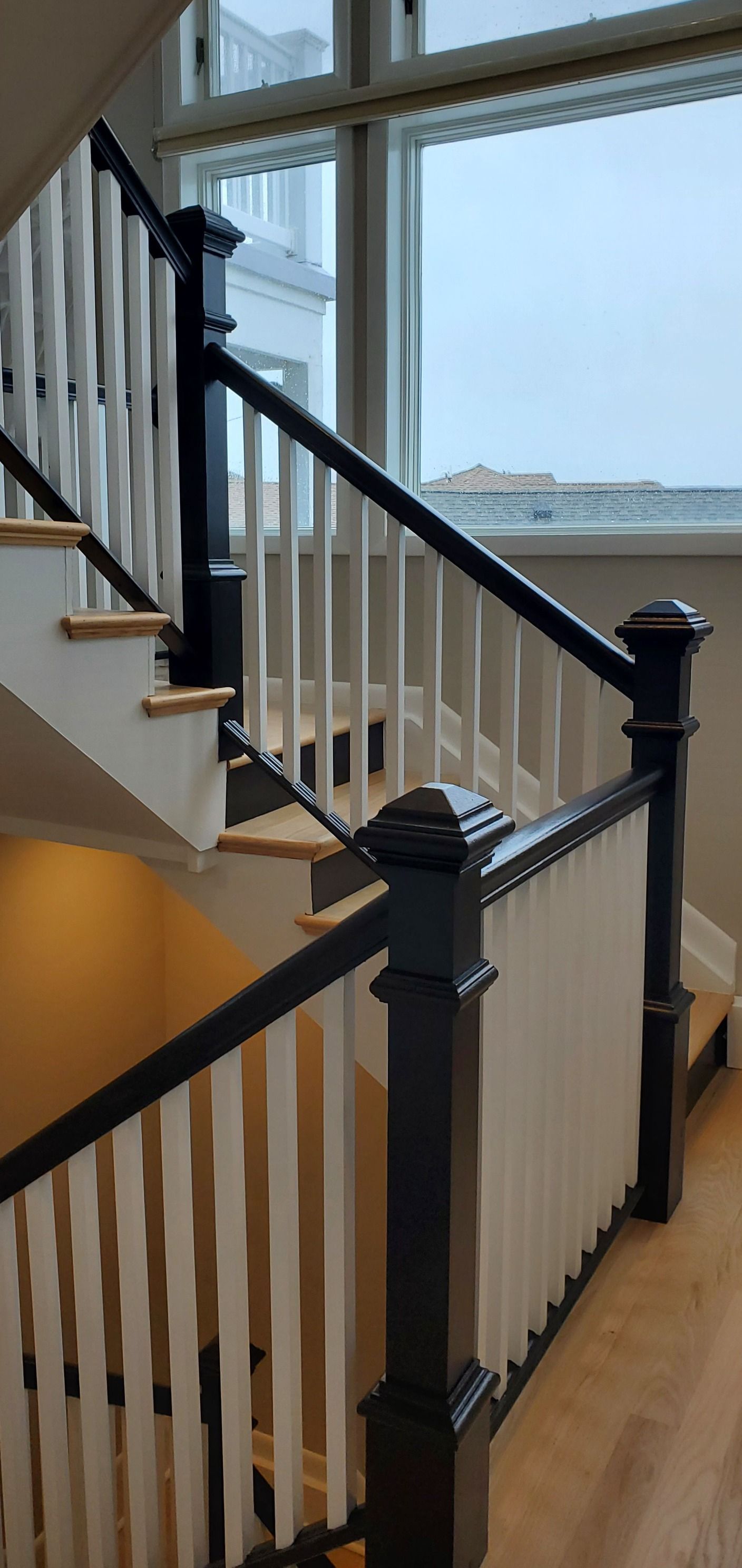 Staircase with white balusters, black handrails and newel posts. Wooden steps. Window in the background.