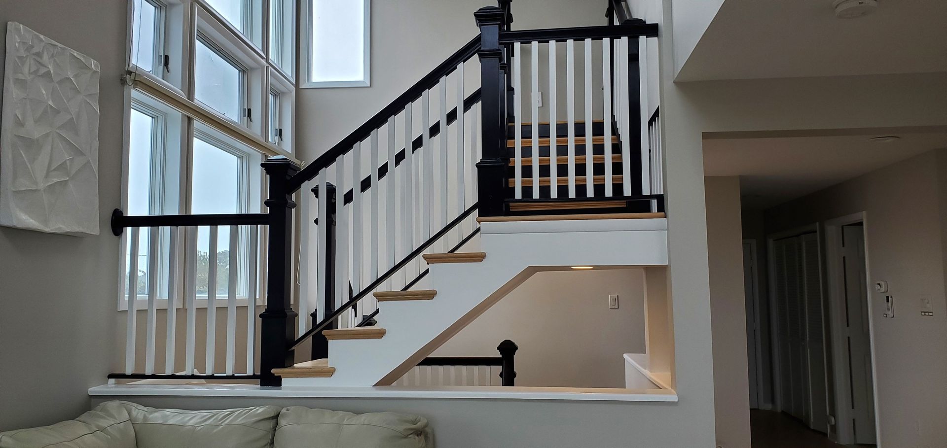 Staircase with black railing and white spindles against a neutral wall. Sunlight streams through tall windows.