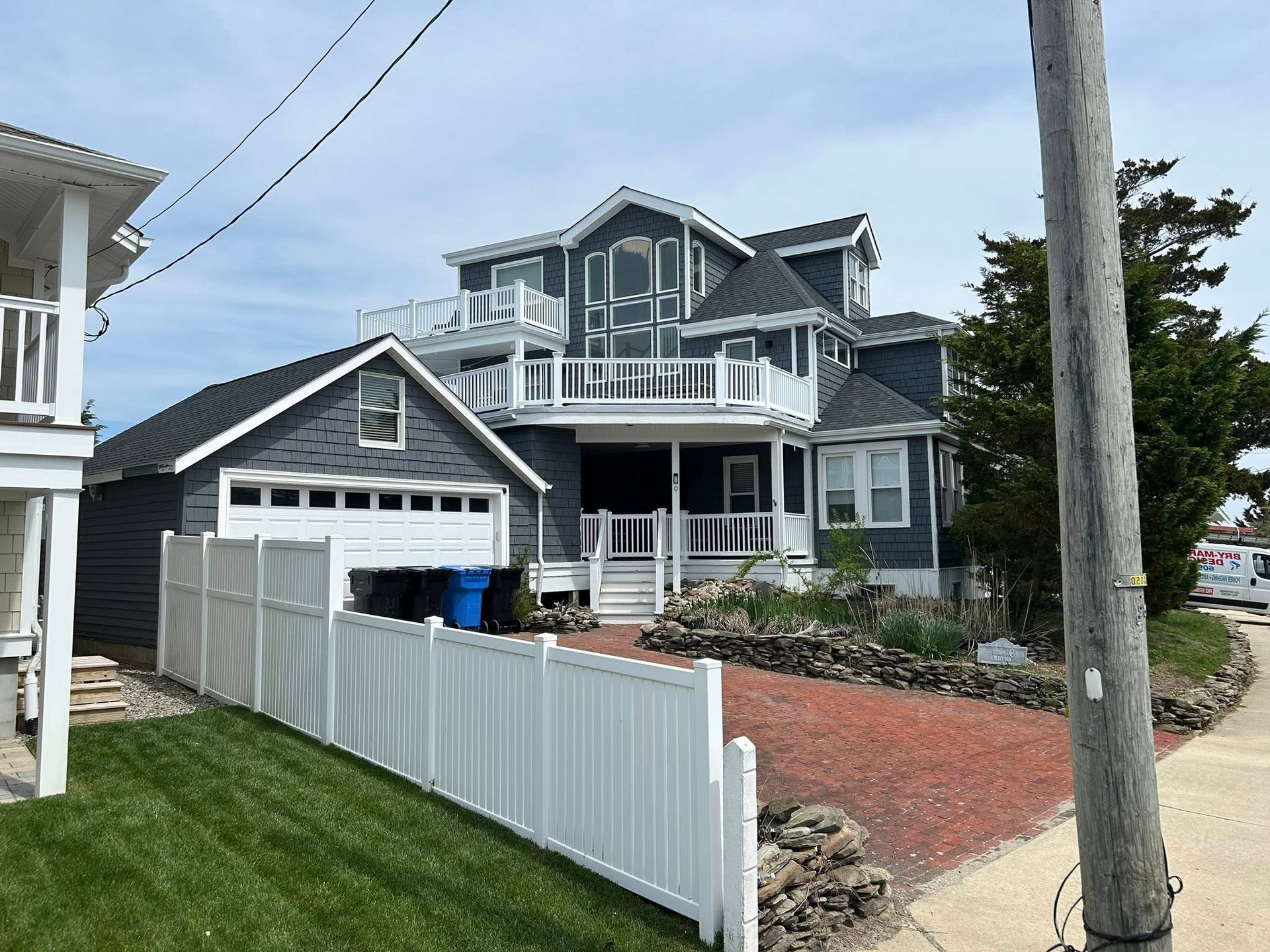 Two-story blue house with white trim, balconies, and garage, behind a white fence and green lawn.