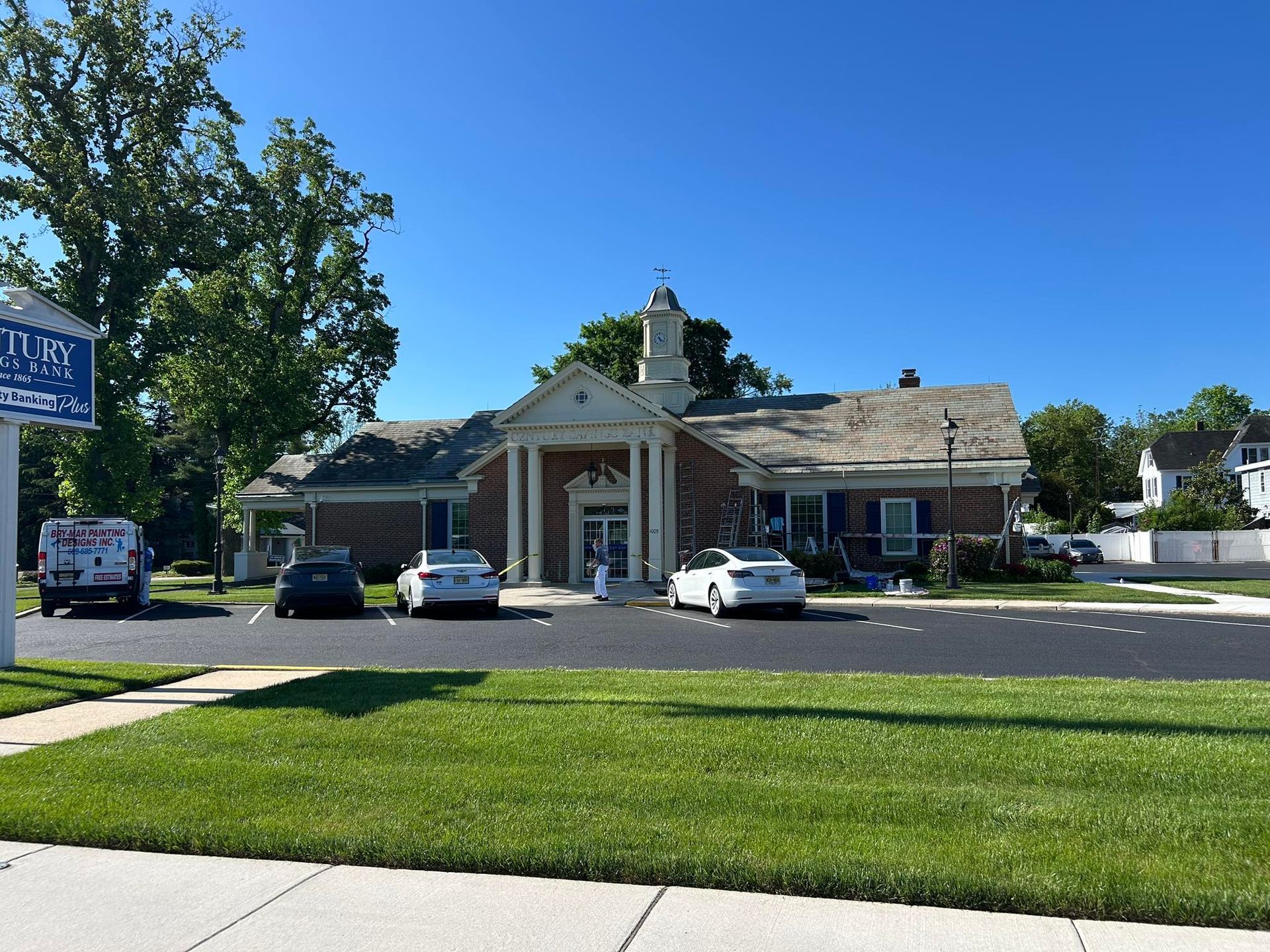 Brick building with columns and a small cupola, parked cars, blue sky, and green grass.