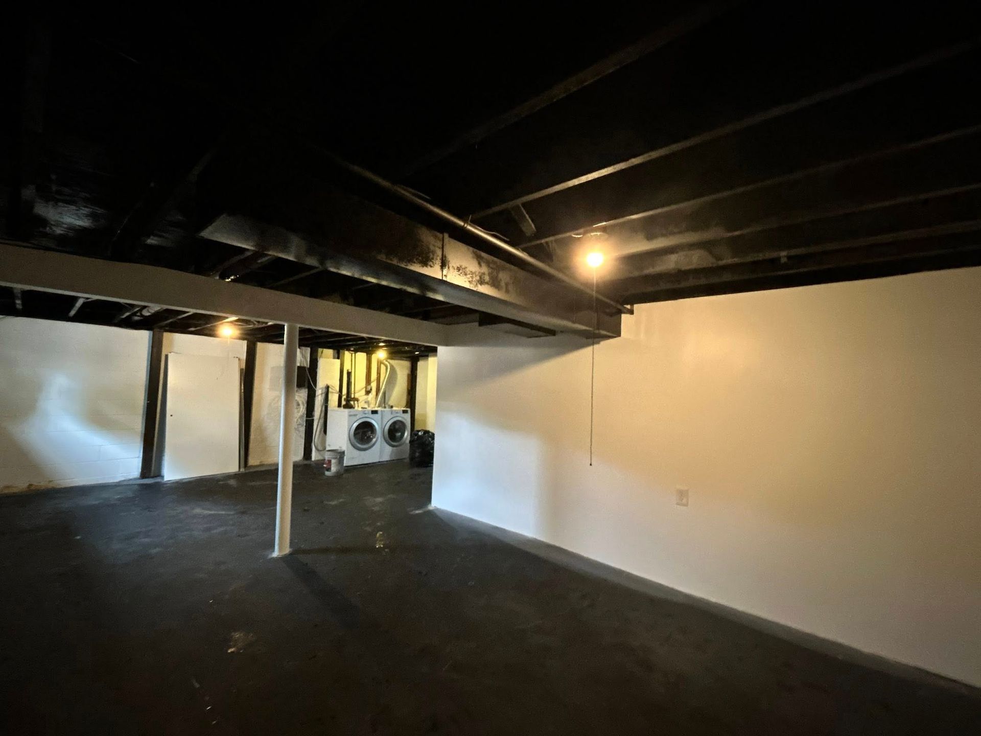 Basement interior with black ceiling and floor, white walls. Washer/dryer visible in the distance.