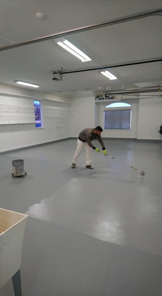 Man rolling paint onto a gray garage floor. A bucket of paint and a utility sink are also visible.