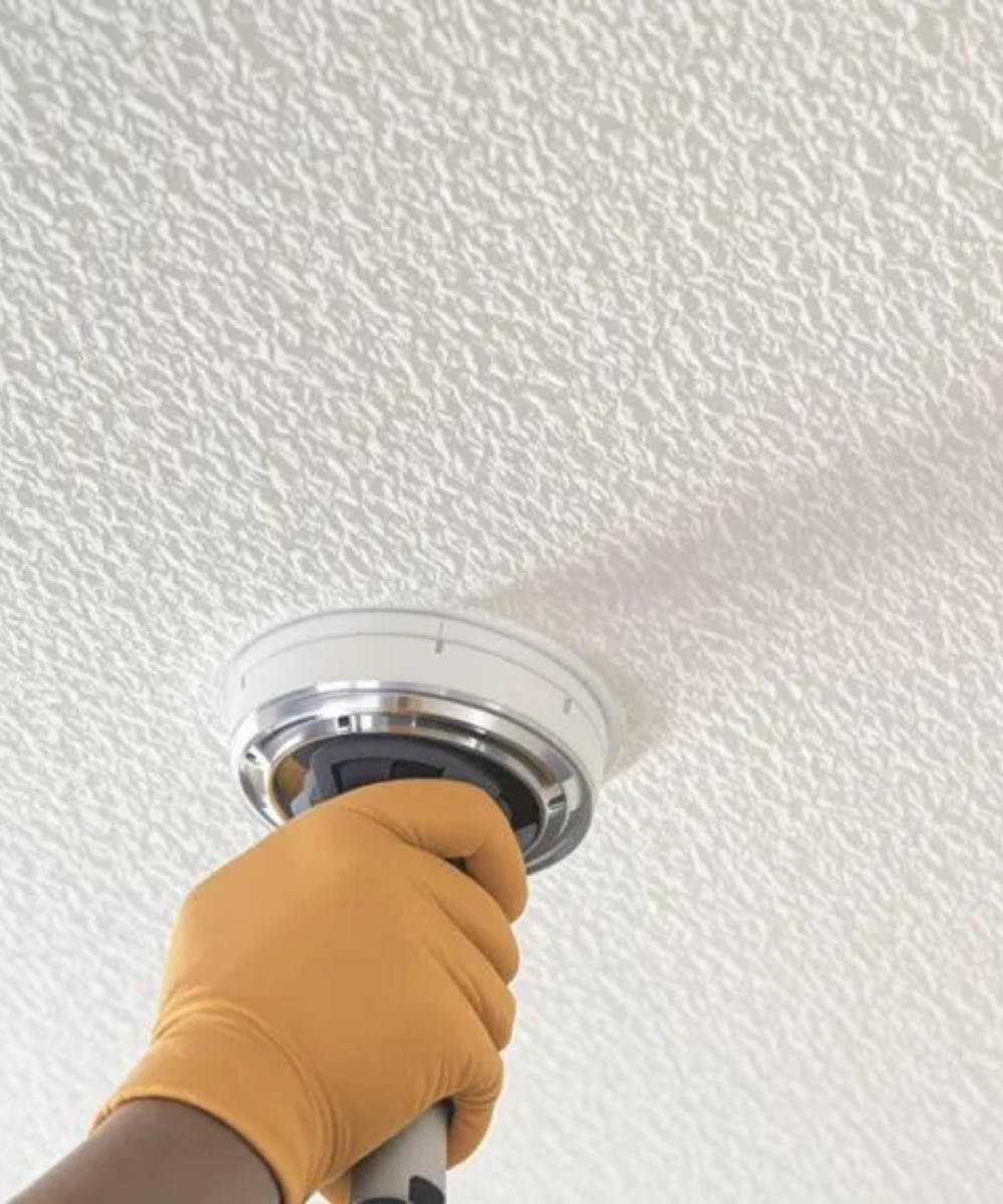 Hand in orange glove holding a tool against a textured white ceiling, likely removing or installing a light fixture.