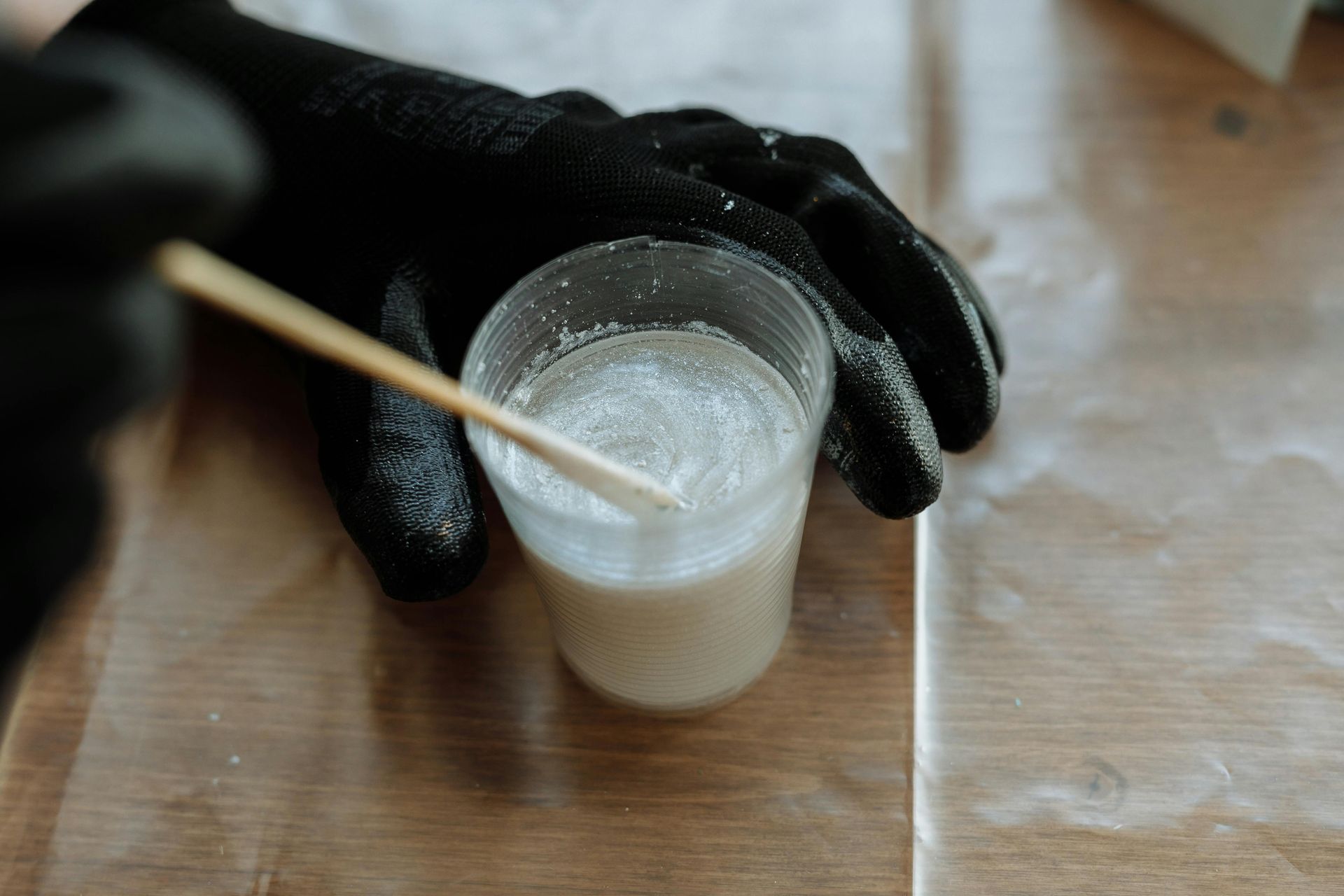 Gloved hand stirring shimmering liquid in a clear cup with a wooden stick on a wooden surface.